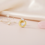 gold open-circle stud earrings with a round center stone and marquise accent on a pink backdrop with a dried flower and a pink stone