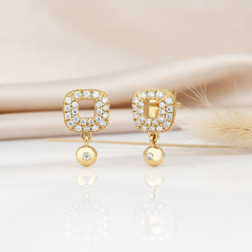 gold open-square stud earrings paved with stones and a dangling gold drop on a cream backdrop with dried grass