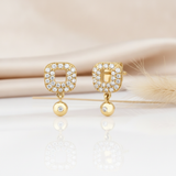 gold open-square stud earrings paved with stones and a dangling gold drop on a cream backdrop with dried grass