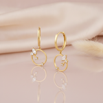 gold huggie hoop earrings with a suspended open ring and a stone cluster on a pink backdrop with dried grass