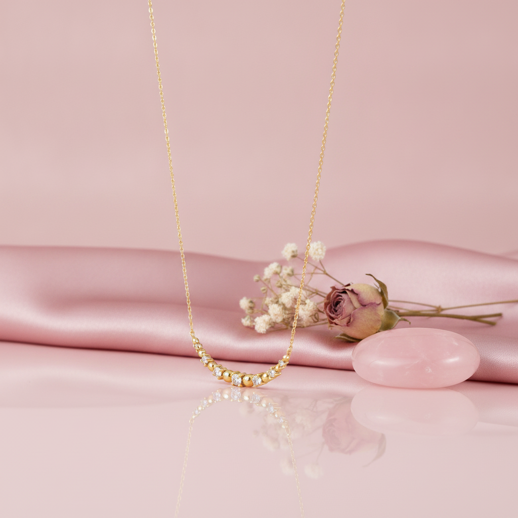 gold necklace with a beaded and stone-set curved arc on a pink backdrop with a dried rose and a rose quartz stone