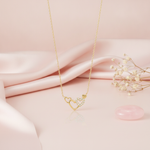 gold pendant necklace with three intertwined heart outlines on a pink backdrop with a dried flower and a rose quartz stone